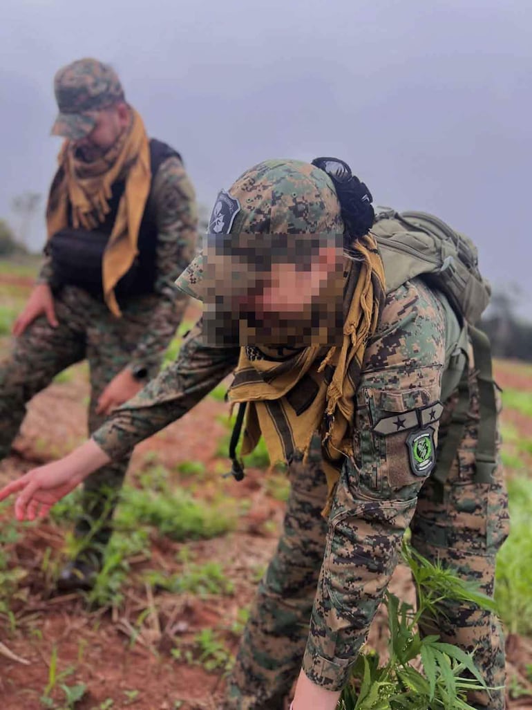Dos soldados vestidos con uniforme camuflado inspeccionan el suelo en un ambiente rural y nublado.