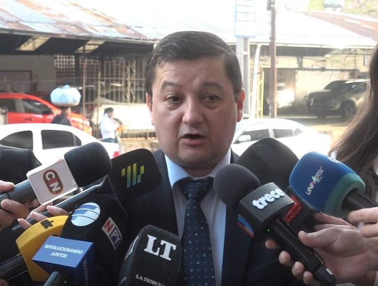 Julio Ortíz en traje oscuro y corbata, centrado ante varios micrófonos mientras periodistas registran sus declaraciones.