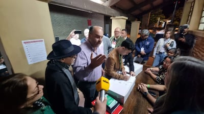 Hombre con sombrero negro gesticula mientras otro firma documentos, rodeados de personas que escuchan atentamente en un ambiente de expectación.