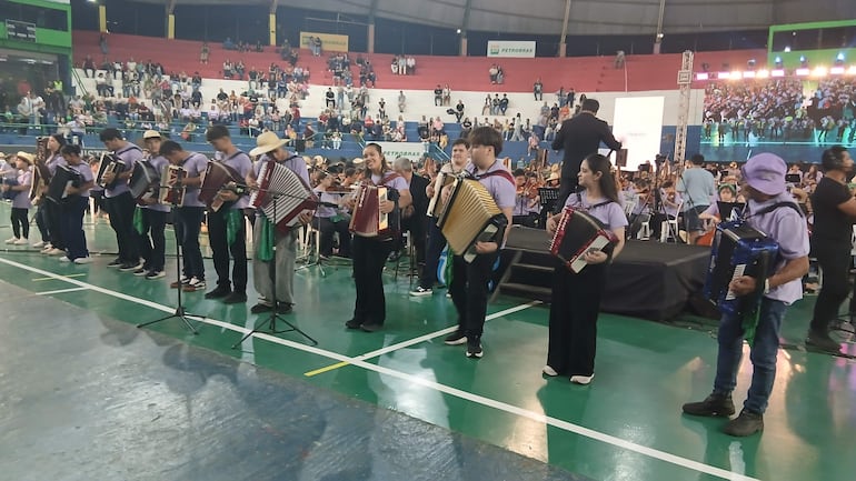 Músicos en camisetas moradas tocan acordeones, mientras un director guía la actuación en un polideportivo lleno de público.