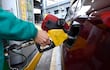 Trabajador con uniforme verde abastece un coche rojo en estación de servicio, manguera amarilla y señales de advertencia visibles.