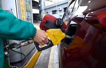Trabajador con uniforme verde abastece un coche rojo en estación de servicio, manguera amarilla y señales de advertencia visibles.
