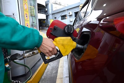 Trabajador con uniforme verde abastece un coche rojo en estación de servicio, manguera amarilla y señales de advertencia visibles.