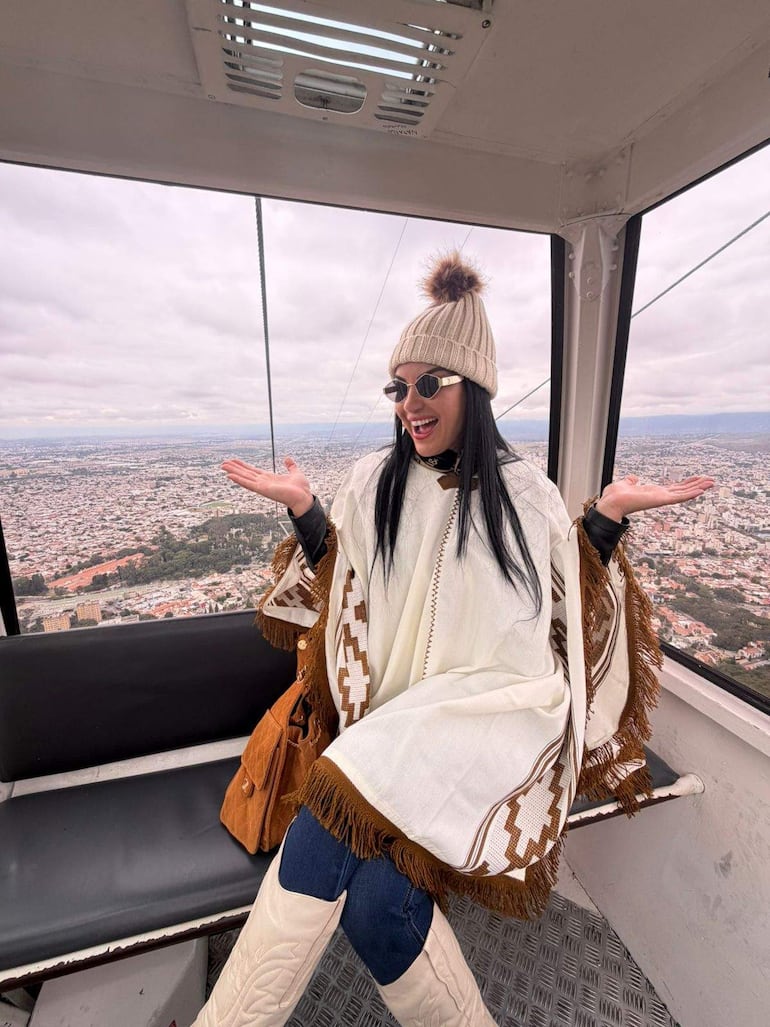 Gloria Vera en teleférico, con abrigo blanco y gorro beige, sonríe mientras gesticula hacia el paisaje urbano.