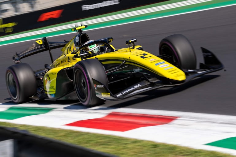 Coche de Fórmula 1 amarillo en carrera, elevado sobre la pista con banderas de colores de fondo.