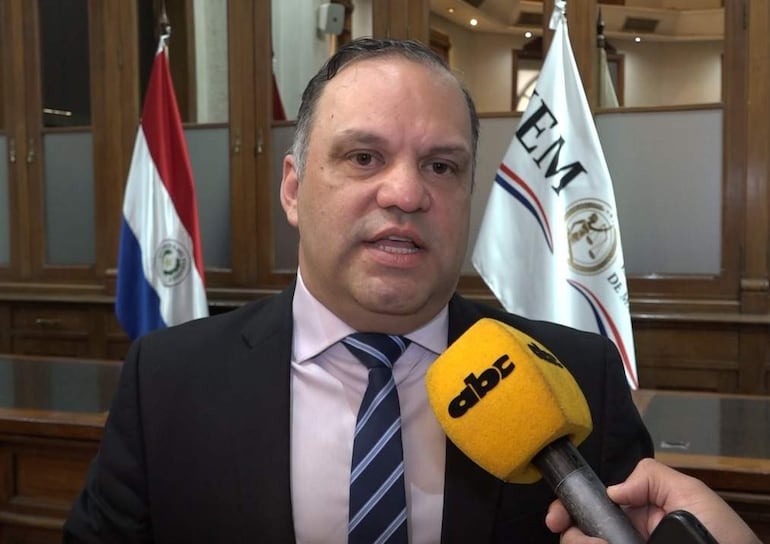 Mario Varela en traje oscuro, con expresión seria, habla con un periodista en un entorno formal, rodeado de banderas.
