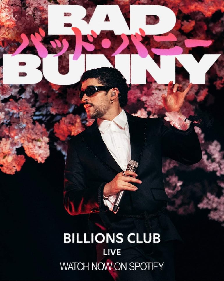 Bad Bunny con traje negro y gafas de sol, sosteniendo un micrófono frente a un fondo de flores rosadas.