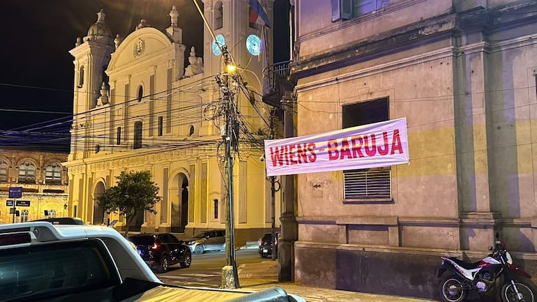 Pasacalle grande con inscripción ‘WIENS-BARUJA’ en letras rojas en entorno urbano nocturno. Fondo con cables eléctricos y luz de calle.