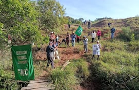 Grupo de productores con banderas verdes y cartel en mano, rodeados de vegetación y un camino de madera.