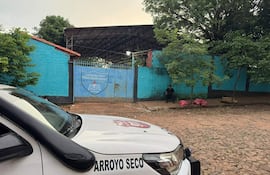 Hombre sentado en silla, rodeado de árboles frente al Colegio Nacional Rosa Agustina Farina, con camioneta policial visible.