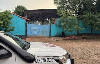 Hombre sentado en silla, rodeado de árboles frente al Colegio Nacional Rosa Agustina Farina, con camioneta policial visible.