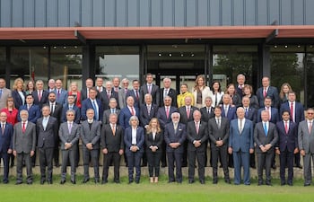 Grupo de 50 personas, en su mayoría vestidas de traje, posan en un ambiente exterior formal con un edificio moderno de fondo.