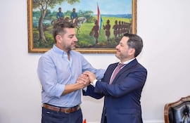 Alliana y Latorre se estrechan las manos, luciendo expresiones amigables. Alliana lleva camisa clara, Latorre un traje oscuro con corbata clara.
