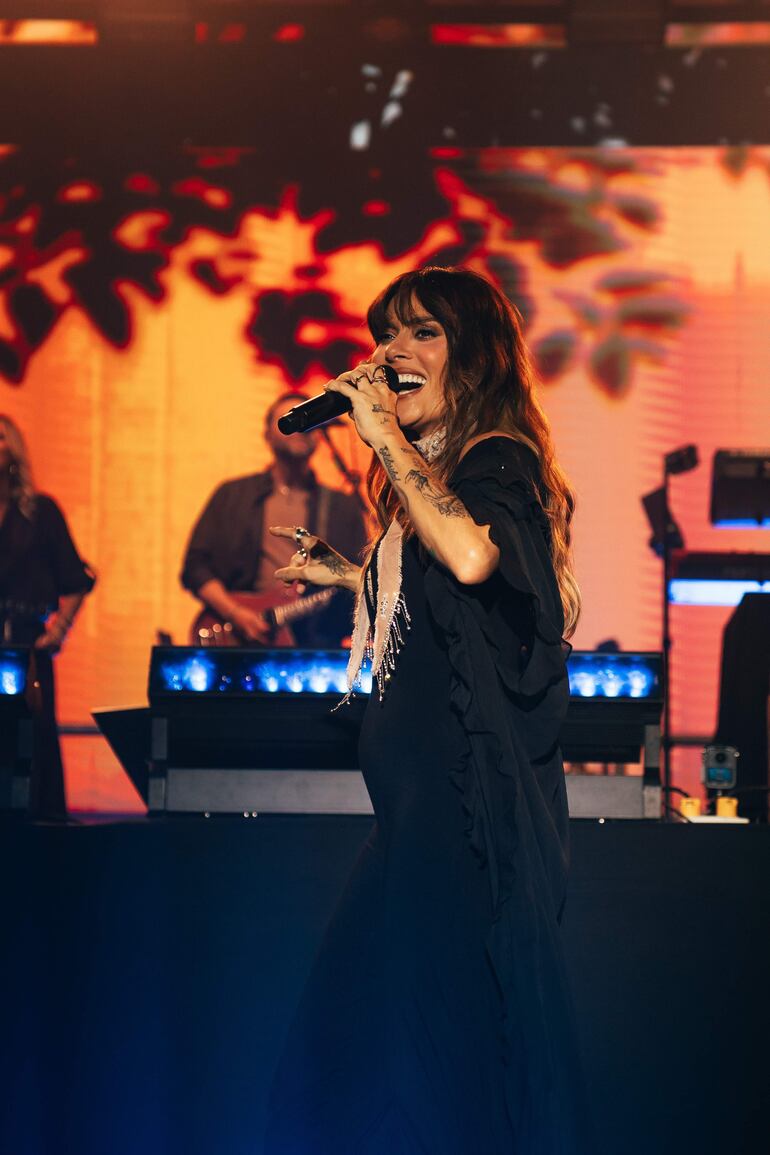 Kany García sonríe en el escenario, sosteniendo un micrófono, vestida de negro con detalles decorativos.