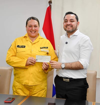 Mujer con uniforme amarillo y hombre con camisa blanca sonríen mientras sostienen un cheque, con una bandera de Paraguay de fondo.
