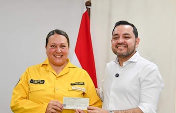 Mujer con uniforme amarillo y hombre con camisa blanca sonríen mientras sostienen un cheque, con una bandera de Paraguay de fondo.