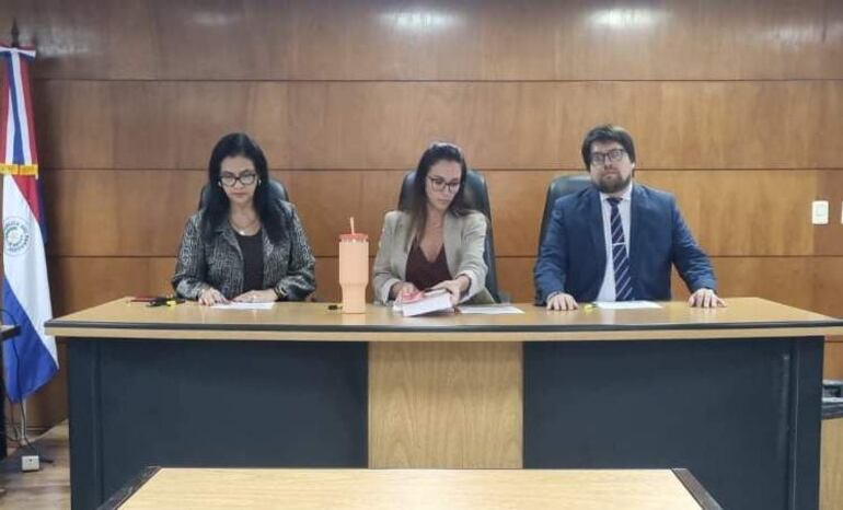 Sonia Sánchez y Adriana Planás revisan documentos sentadas en una mesa, mientras Matías Garcete observa con atención en un ambiente formal.