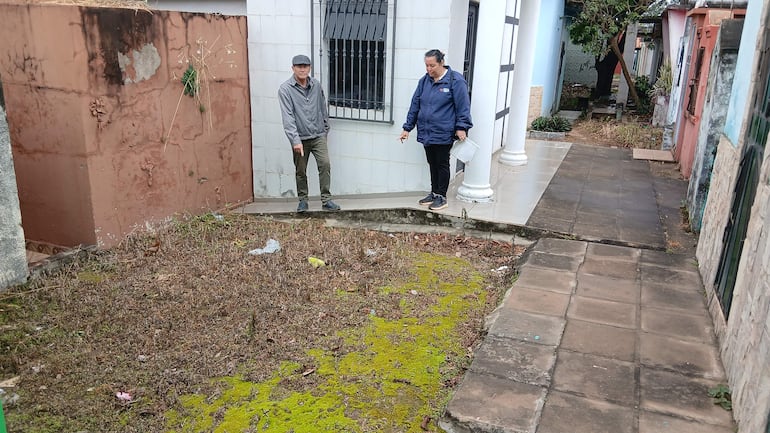 Felicia Ramona Benítez Aguilera en chaqueta azul sostiene un recipiente mientras un hombre con gorra señala el terreno irregular del cementerio.