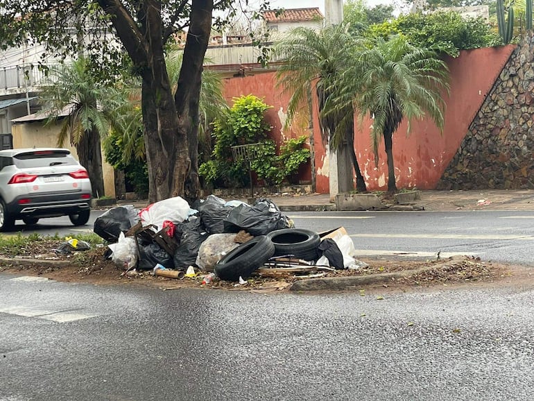 Montículo de basura acumulada en Tembetary, con casas y palmeras al fondo, un vehículo gris circulando.