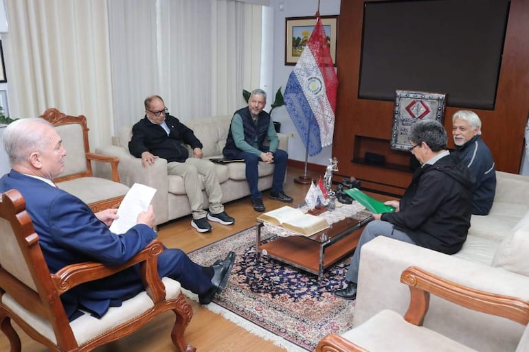 Hombre de traje oscuro sostiene documentos mientras otros tres hombres escuchan en una sala con bandera de Paraguay de fondo.