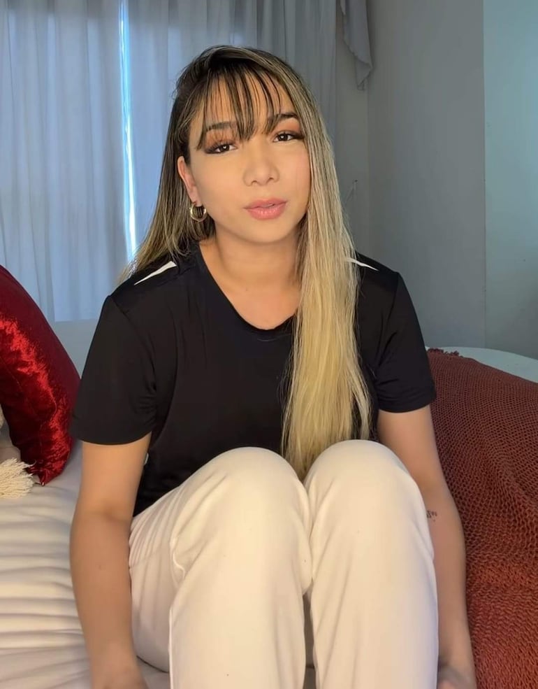 Marilina Bogado sentada en una cama, con cabello lacio y flequillo, vestida con camiseta negra y pantalones blancos, en un ambiente iluminado.