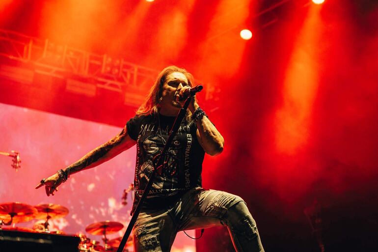 Cantante masculino con cabello largo y barba, vestido de negro, sostiene un micrófono enérgicamente bajo luces rojas intensas.