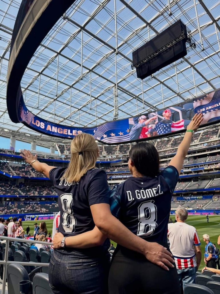 Dos mujeres de espaldas, una con el brazo alzado, vistiendo camisetas oscuras con el apellido 'Gómez' y el número 8, en un estadio con ambiente festivo.