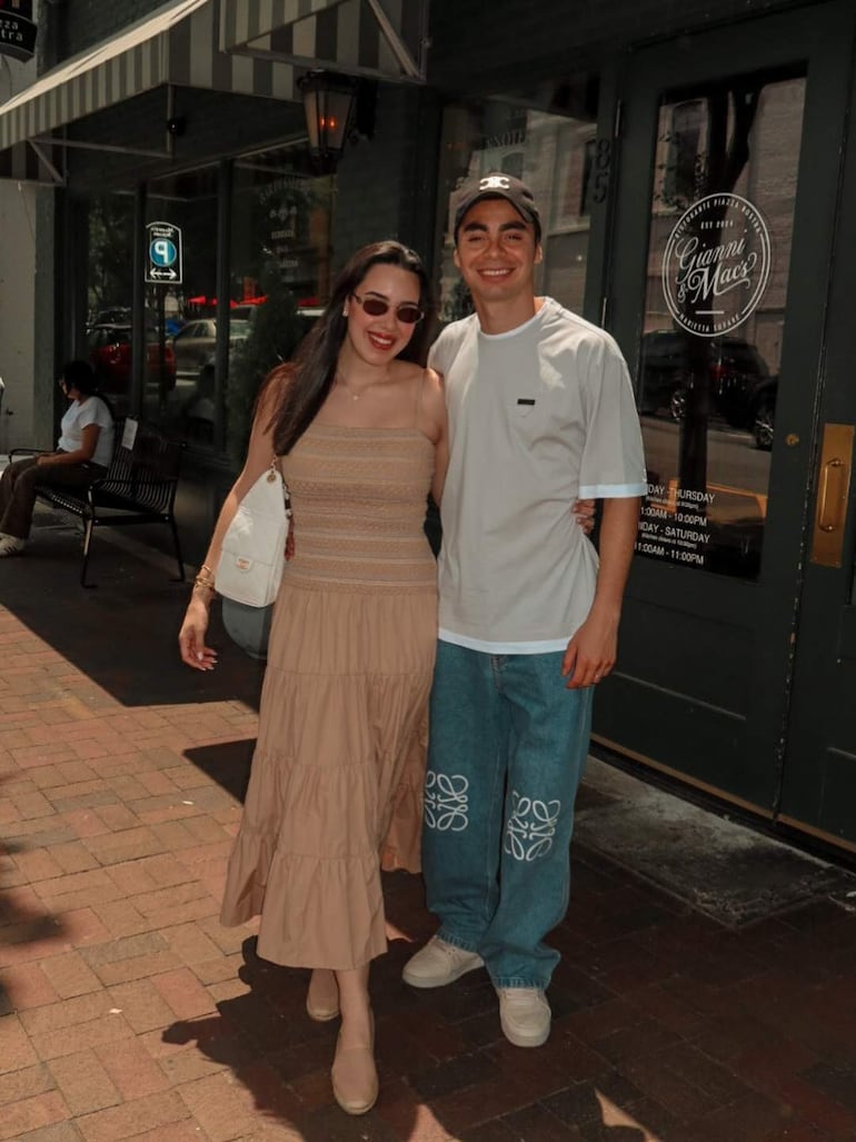 Miguel Almirón con camiseta blanca y Alexia Notto en vestido beige y gafas de sol, sonríen en un ambiente urbano junto a un restaurante.