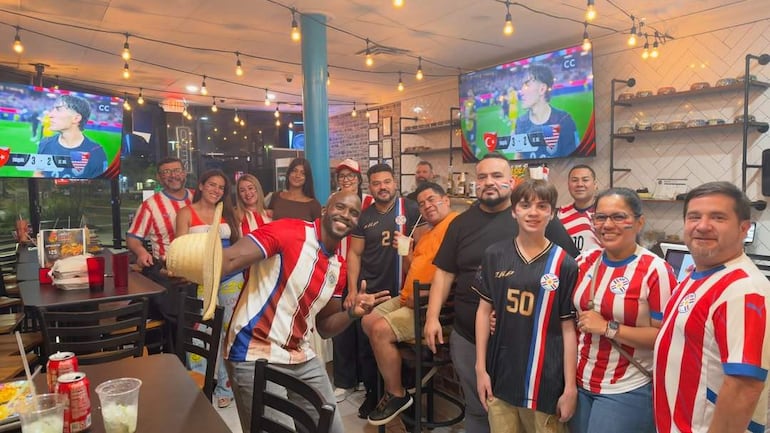 Hinchas en bar, felices y animados, visten camisetas paraguayas mientras observan un partido en pantallas luminosas.