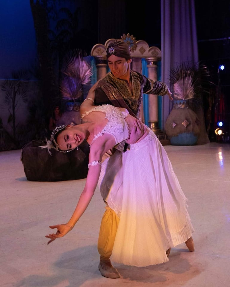 Bailarín en traje oscuro sostiene a bailarina en vestido blanco con diadema en una actuación dramática sobre el escenario.