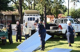 Hombre en uniforme policial agachado sobre chapas metálicas mientras otros observan. Ambiente tranquilo y árboles al fondo.