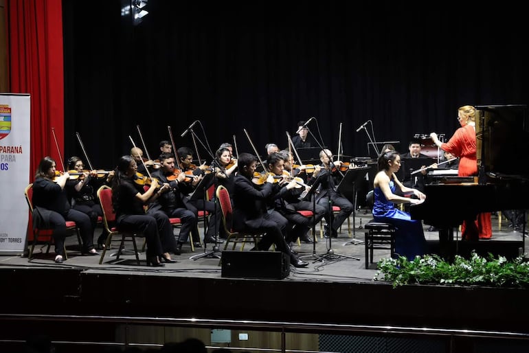 Pianista en vestido azul y director en rojo dirigen a la orquesta con músicos en ropa negra, en un escenario con telón rojo.