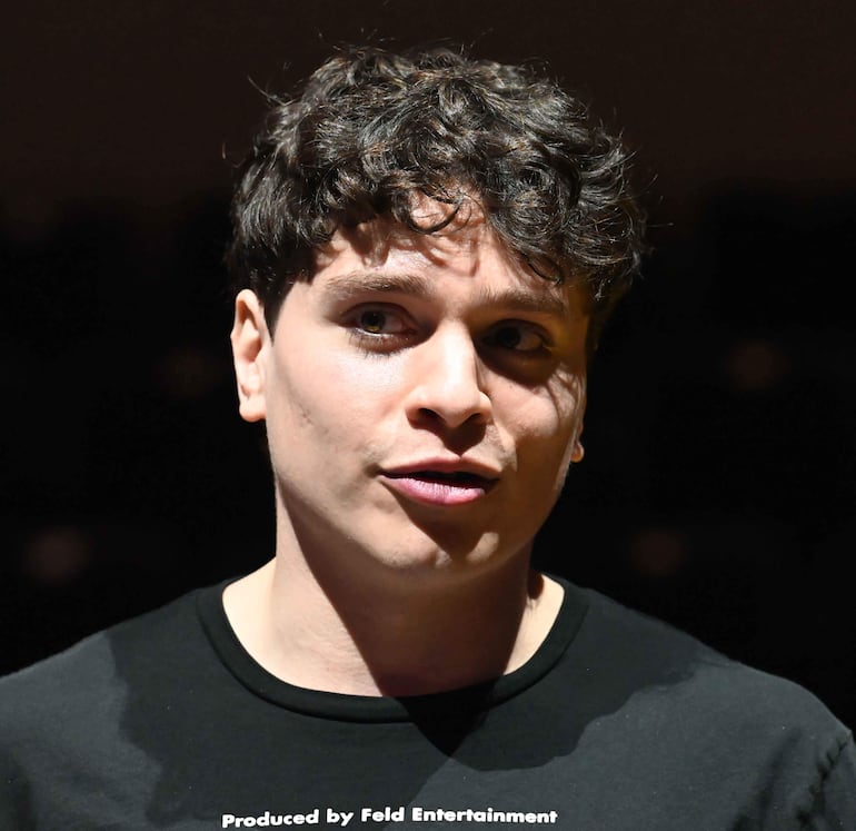Joven con cabello rizado y oscuro, vistiendo camiseta negra, habla en un ambiente oscuro con luces suaves.