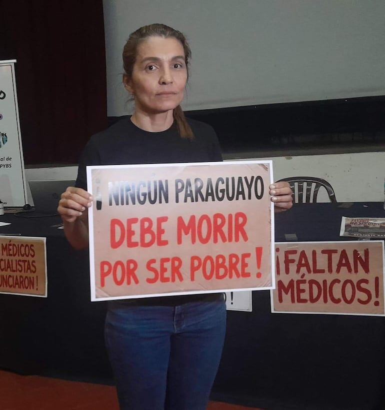 Mujer de pie con camiseta negra y jeans, sosteniendo cartel que dice '¡Ningún paraguayo debe morir por ser pobre!'.