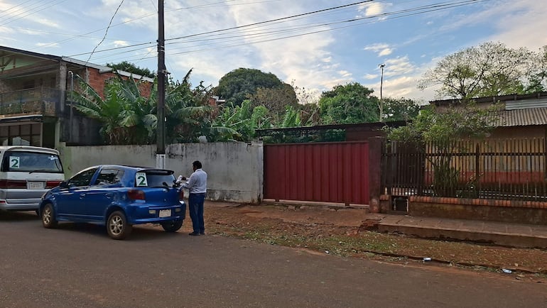 Un hombre de pie junto a un auto azul, sosteniendo un dispositivo, en una escena urbana con vehículos y vegetación alrededor.