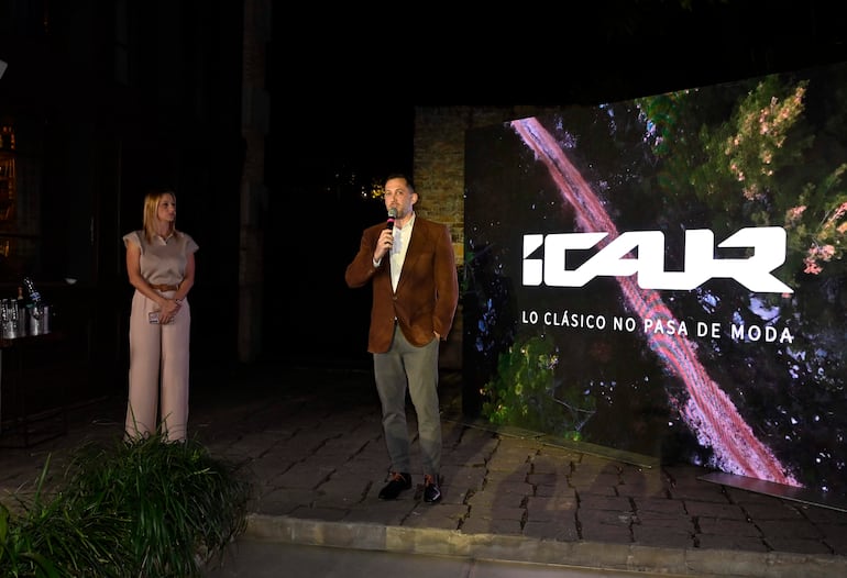 Mujer en conjunto claro escucha mientras hombre en blazer marrón habla con micrófono frente a logo iluminado de ICAUR.