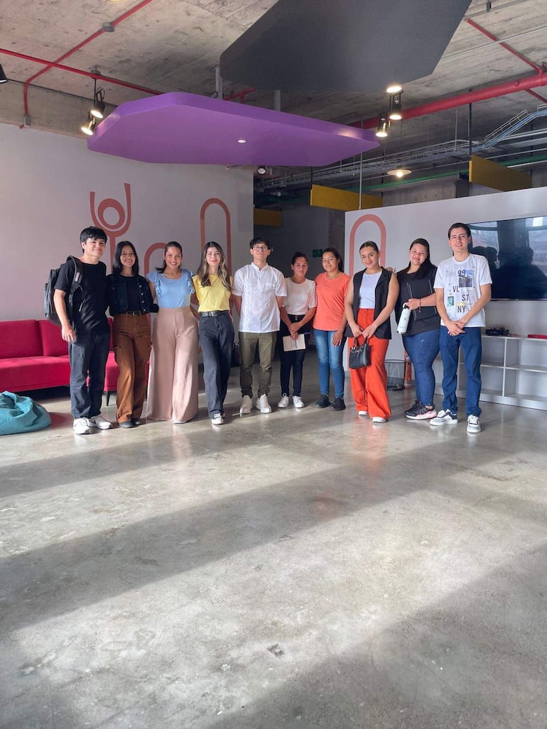 Grupo de diez estudiantes sonrientes, siete mujeres y tres hombres, posando con mochilas y botellas en un espacio moderno con sofá rojo.