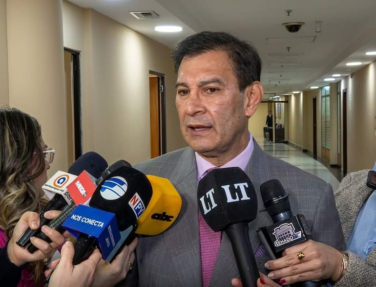 Silvio Ovelar en traje gris y corbata rosa responde a periodistas, rodeado de micrófonos en un pasillo con luz artificial.