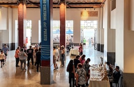 Un hombre sentado cerca de una mesa con productos artesanales, mientras otras personas conversan y observan, en un ambiente vibrante.