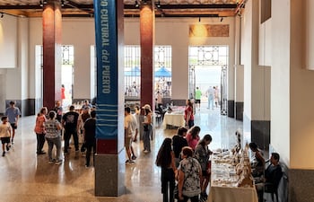 Un hombre sentado cerca de una mesa con productos artesanales, mientras otras personas conversan y observan, en un ambiente vibrante.