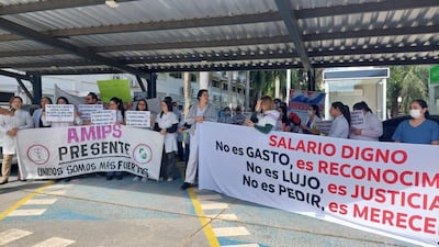 Grupo de médicos en batas y camisetas claras sostiene pancartas en protesta, bajo una estructura techada.