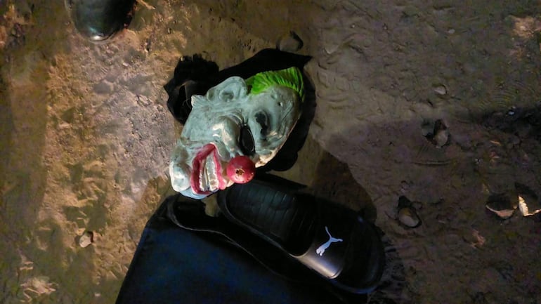 Máscara de payaso con cabello verde y sandalia negra en el suelo, junto a tierra y piedras. Sin personas visibles.