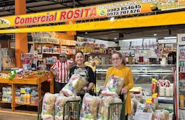 Dos mujeres sonrientes con bolsas de compras en el mercado, rodeadas de productos alimenticios y un hombre observando.