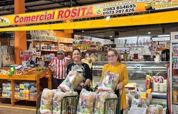 Dos mujeres sonrientes con bolsas de compras en el mercado, rodeadas de productos alimenticios y un hombre observando.