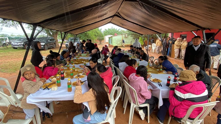 Grupo de personas en mesa bajo carpa, compartiendo comida y bebida, con policías en uniforme de fondo.