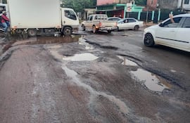Escena de tráfico con un camión blanco, automóvil gris y baches llenos de agua en un entorno urbano con edificios coloridos.
