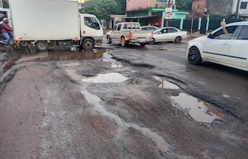 Escena de tráfico con un camión blanco, automóvil gris y baches llenos de agua en un entorno urbano con edificios coloridos.