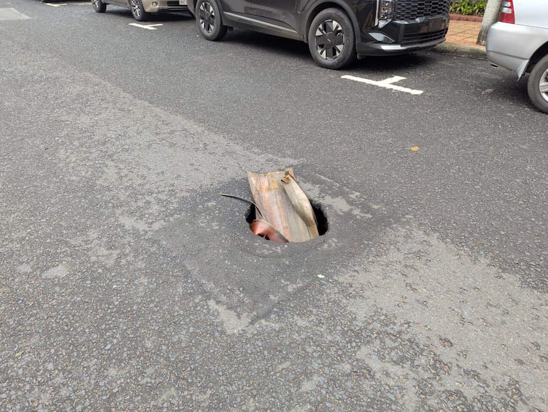 Bache profundo en la carretera con un trozo de metal visible, rodeado de vehículos estacionados.