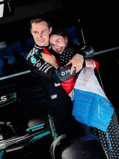 Joshua Duerksen en overol negro y Anneke Rahn en vestido multicolor, sonriendo y abrazándose en un taller de automovilismo.