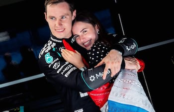 Joshua Duerksen en overol negro y Anneke Rahn en vestido multicolor, sonriendo y abrazándose en un taller de automovilismo.